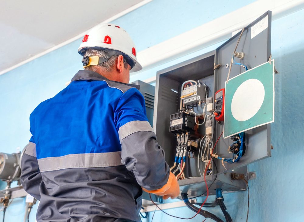 Electrician wearing a hard hat and protective gear inspecting internal wiring and electrical components inside an industrial panel box