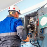 Electrician wearing a hard hat and protective gear inspecting internal wiring and electrical components inside an industrial panel box