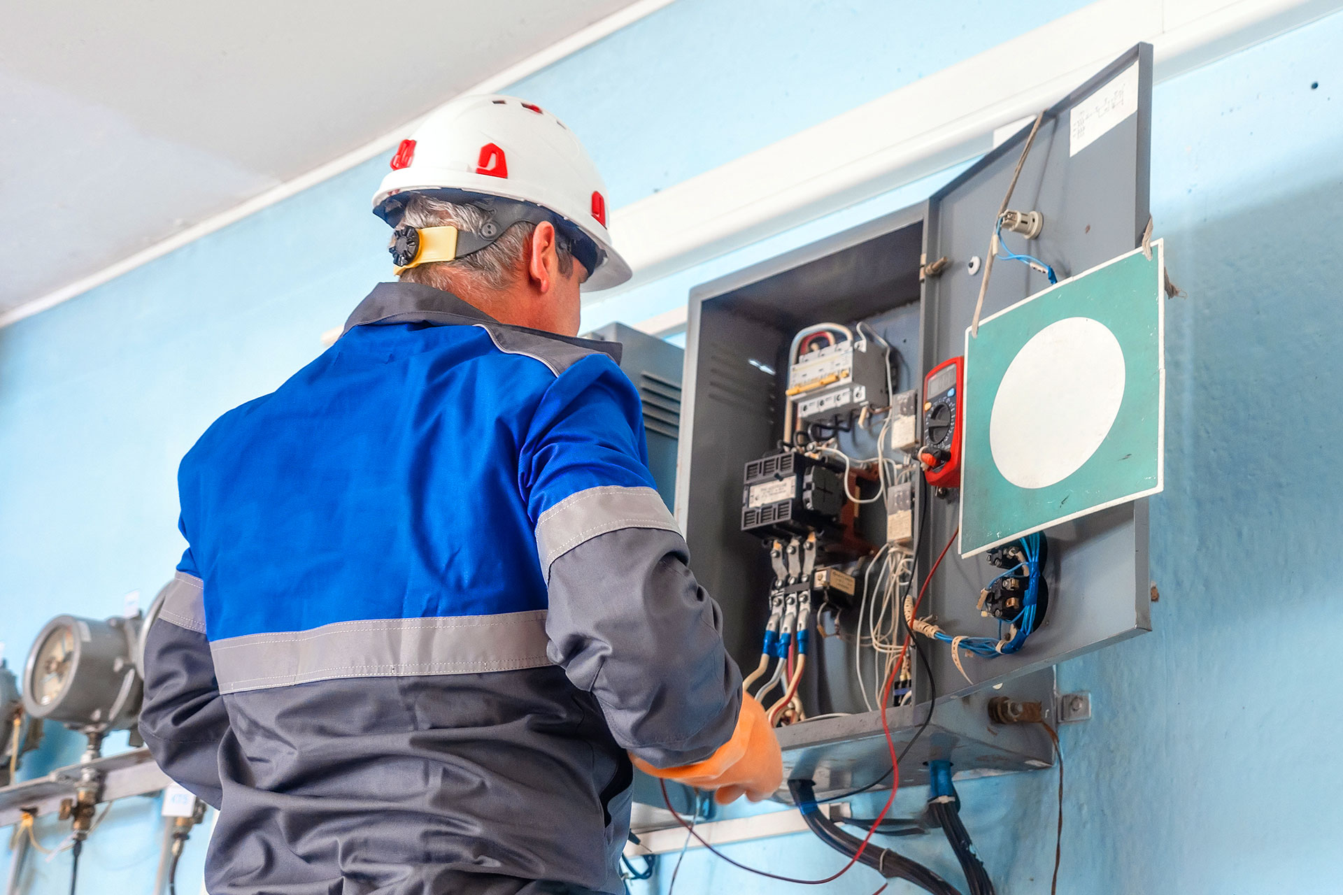 Electrician wearing a hard hat and protective gear inspecting internal wiring and electrical components inside an industrial panel box